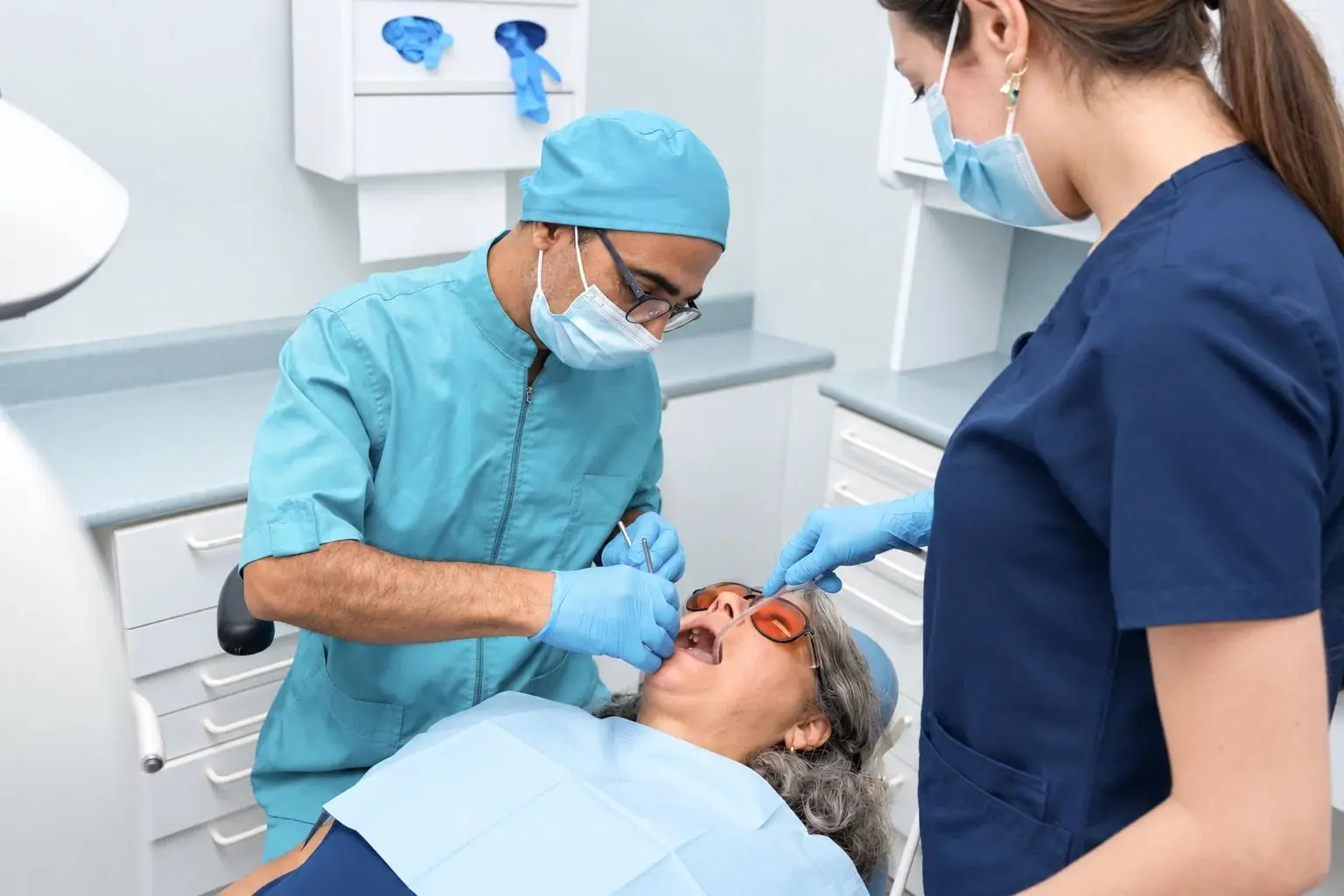 A dentist in a teal cap and mask examines an older patient’s teeth while an assistant in navy scrubs helps