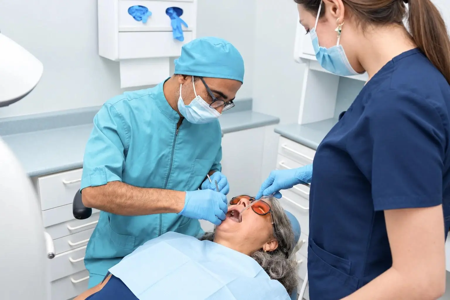 A dentist in a teal cap and mask examines an older patient’s teeth while an assistant in navy scrubs helps