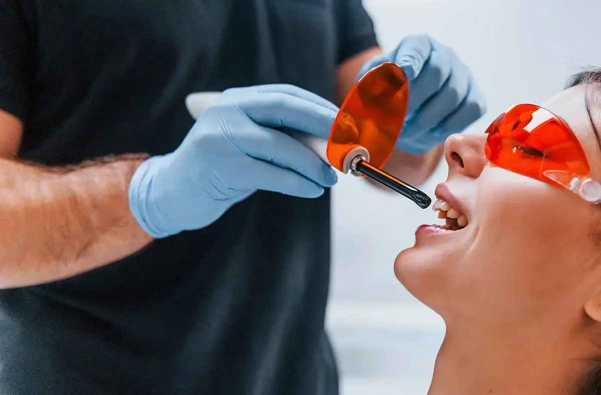 A dental professional, dentist wearing gloves and using a curing light during a dental procedure on a patient wearing protective eyewear.