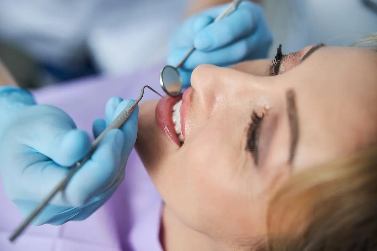 The image shows a close-up of a dental examination. A female patient with blonde hair is lying back in a dental chair with her mouth slightly open, smiling. A dentist or dental hygienist wearing blue gloves is using dental instruments, including a mirror and a probe, to inspect her teeth. The patient appears relaxed, and the setting is a clinical environment. Let me know if you need a more detailed description!