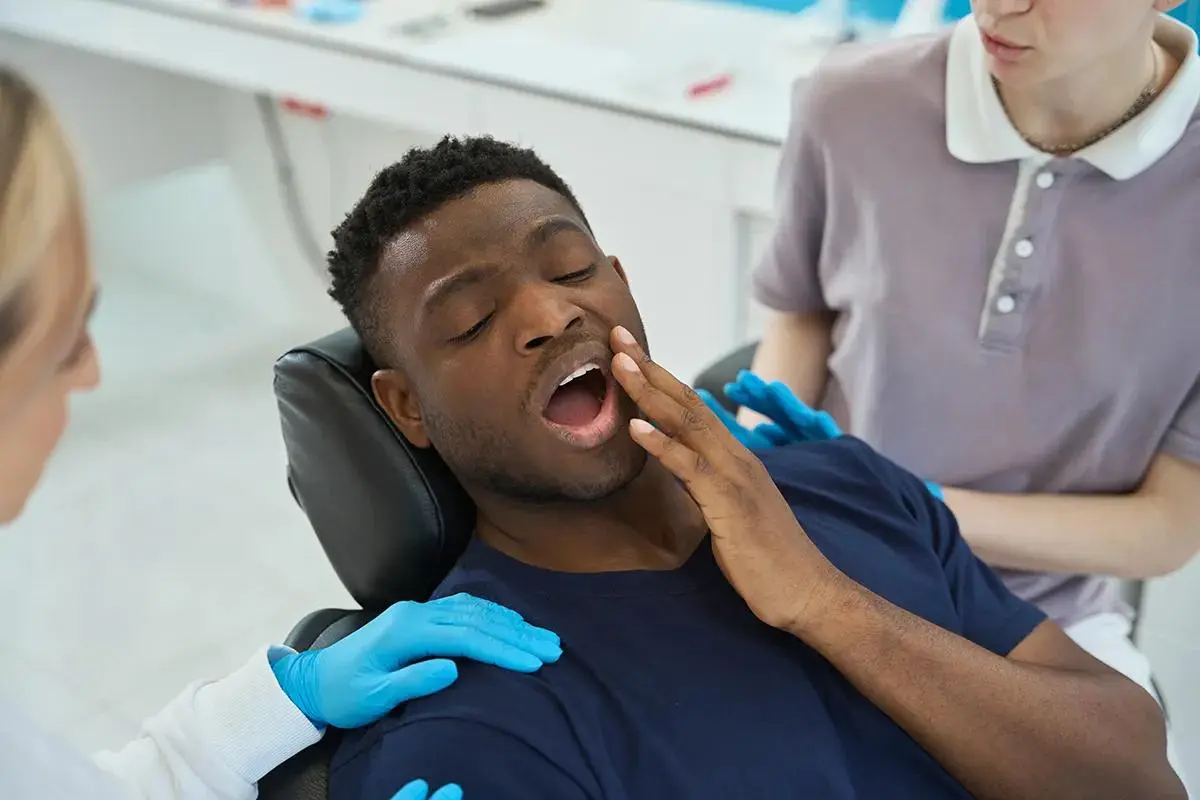 Patient experiencing dental discomfort touching jaw in dental chair with healthcare professionals in blue gloves providing care.