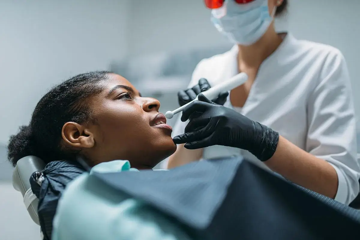 A dentist in protective glasses examines a reclining patient's teeth