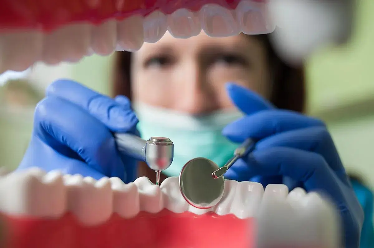 Close-up view through open mouth model showing dentist in blue gloves and mask holding dental mirror and probe instruments, demonstrating dental examination procedure from patient's perspective