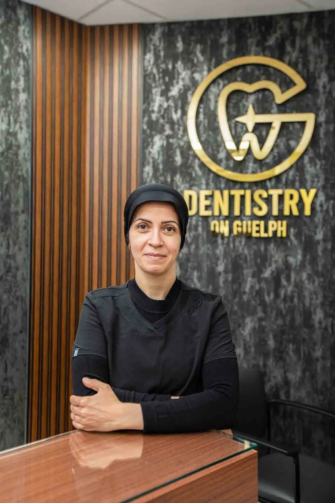 Dr. Mozhgan in black scrubs, arms folded on the reception counter in front of the gold Dentistry on Guelph logo
