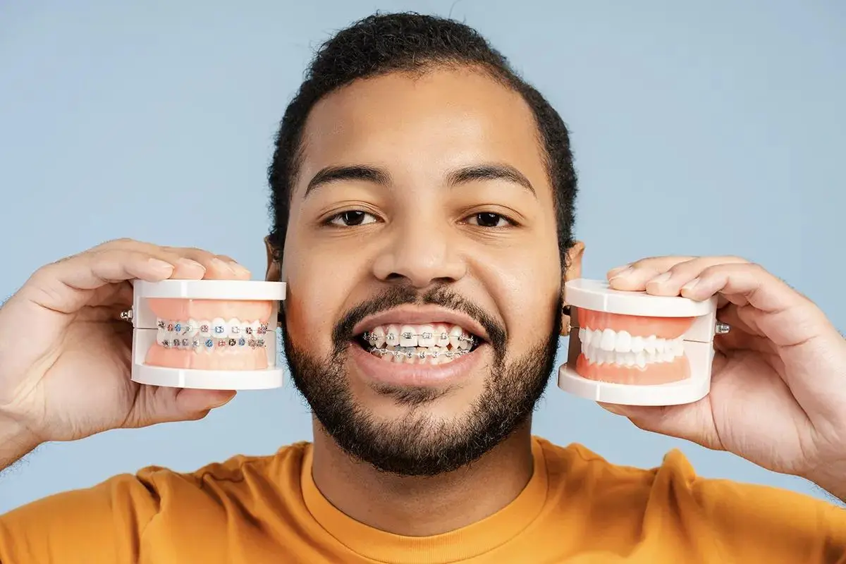 A smiling patient wearing traditional braces while holding dental models showing braces and natural teeth alignments