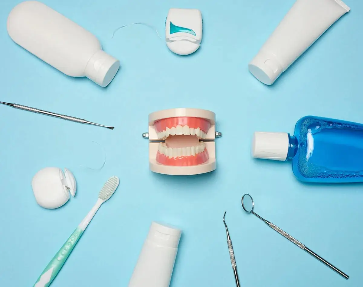 Dental hygiene products and tools arranged around jaw model on blue surface - toothbrush, floss, mouthwash, toothpaste tubes and dental examination instruments