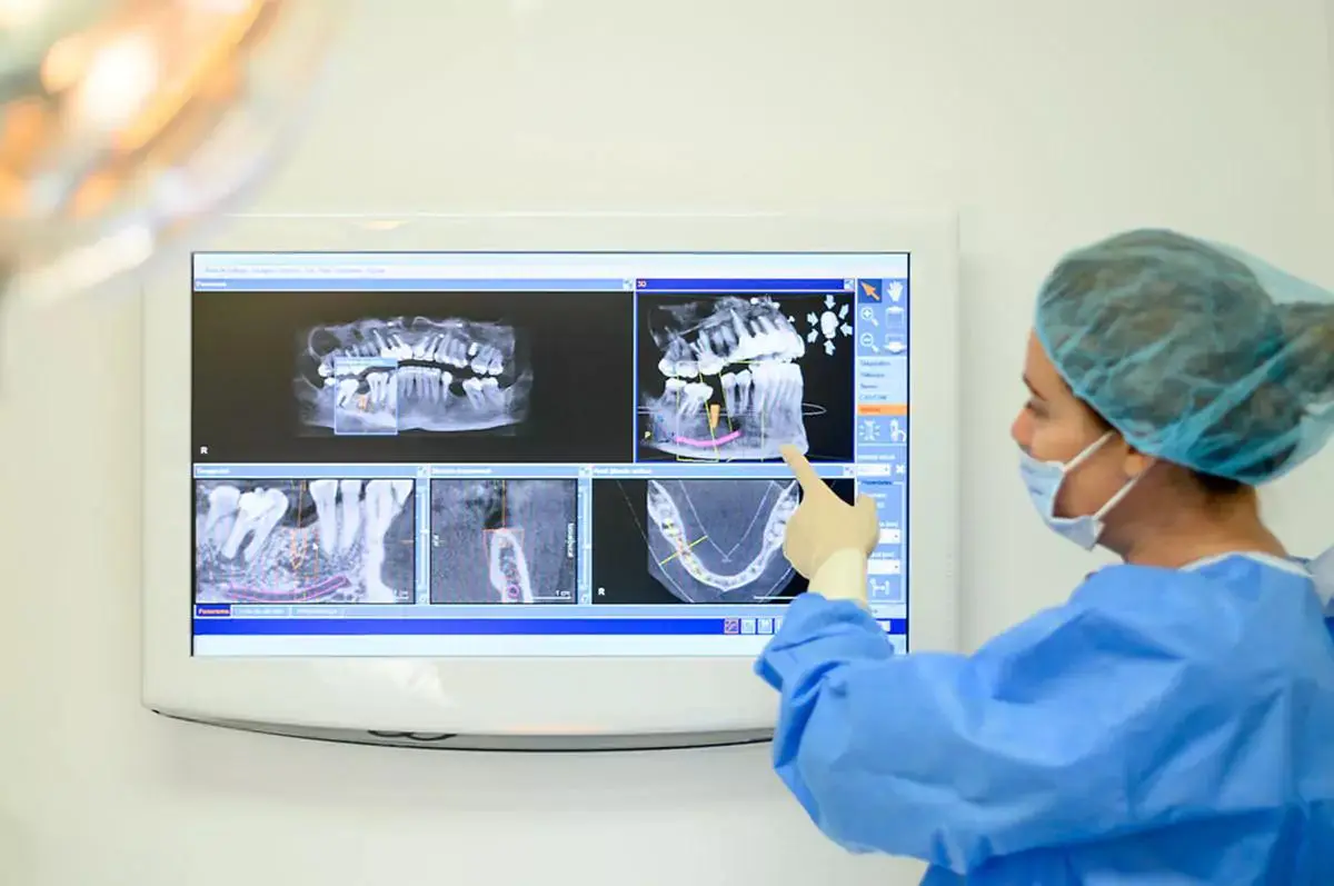 A dental professional in surgical attire examining detailed dental x-rays and scans on a monitor for implant planning.