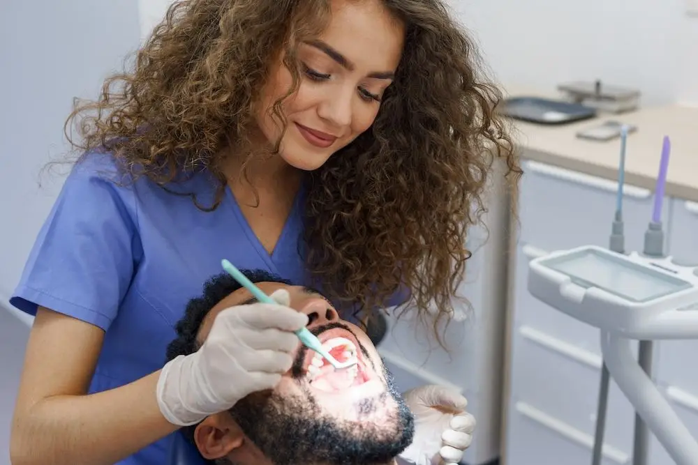A young woman dentist with curly hair, wearing blue scrubs and gloves, is performing a dental examination on a male patient. She is holding a dental instrument and a mirror, focusing on the patient's open mouth. The dental office is equipped with various tools and equipment in the background, creating a professional and clean environment.