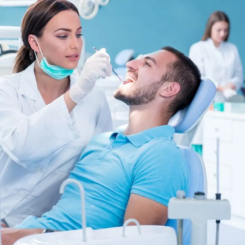 A dentist examines a smiling man's teeth while an assistant works in the background