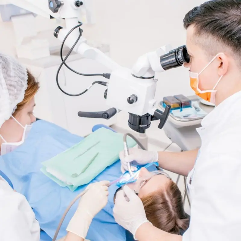 A dentist uses a dental microscope during treatment while an assistant helps