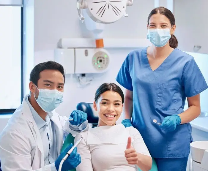 Two dental professionals in masks and a smiling patient giving a thumbs up in a dental chair