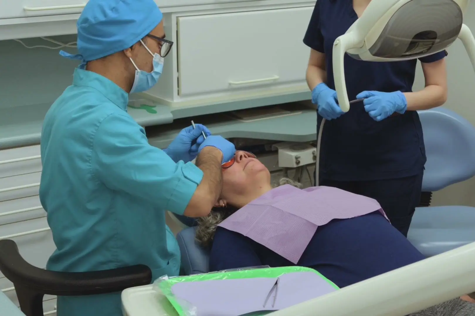 A masked dentist treats a reclining patient wearing protective glasses while a dental assistant stands beside the chair