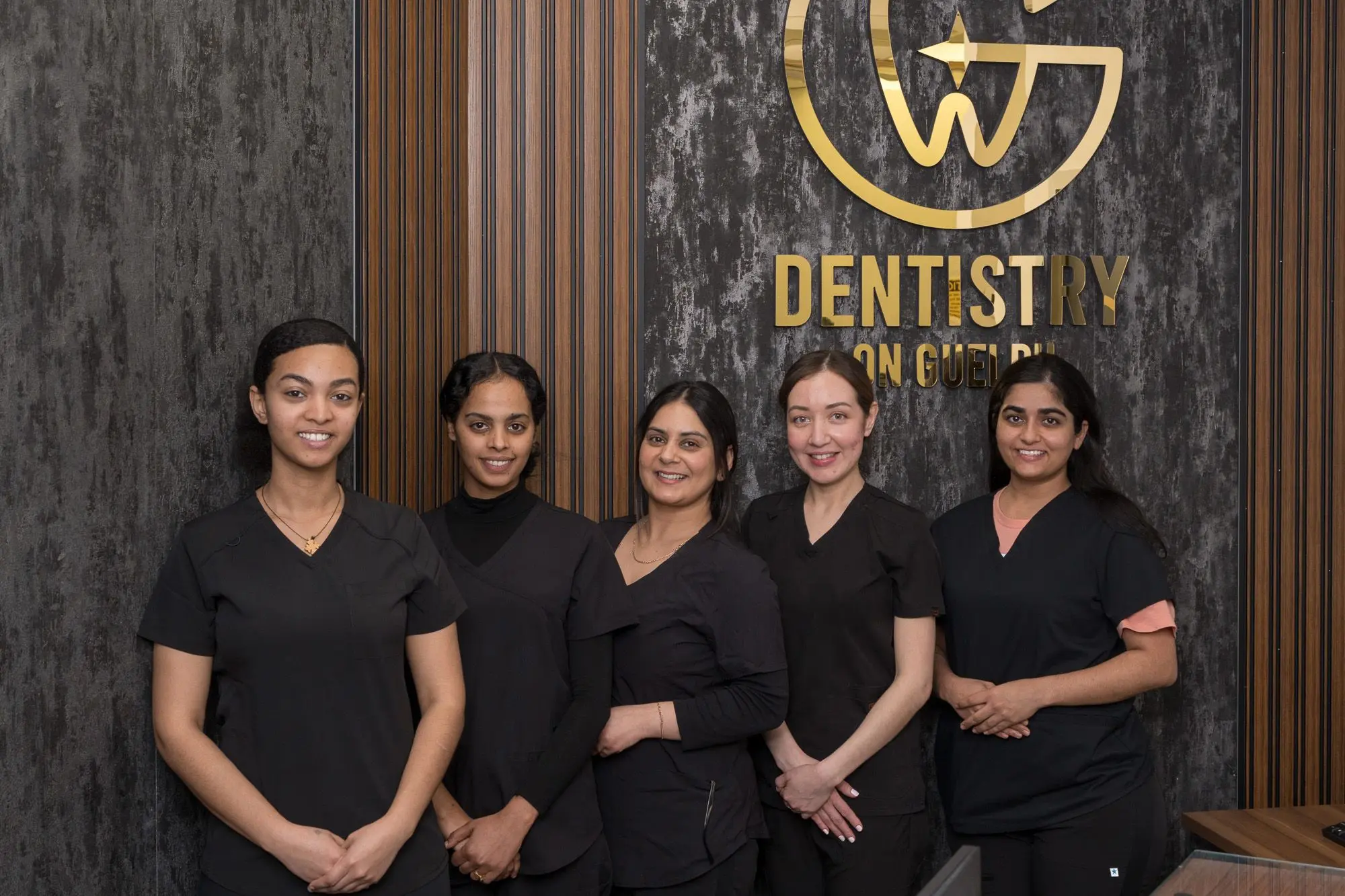 Five Dentistry on Guelph team members in black scrubs stand together in front of the gold clinic logo
