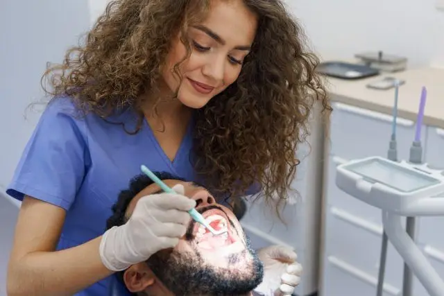 A young woman dentist with curly hair, wearing blue scrubs and gloves, is performing a dental examination on a male patient. She is holding a dental instrument and a mirror, focusing on the patient's open mouth. The dental office is equipped with various tools and equipment in the background, creating a professional and clean environment.