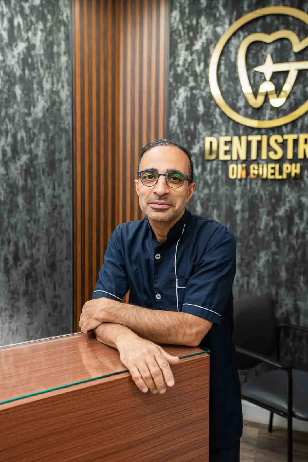 Dr. Reza in a navy dental uniform, leaning on the reception counter in front of the gold Dentistry on Guelph logo