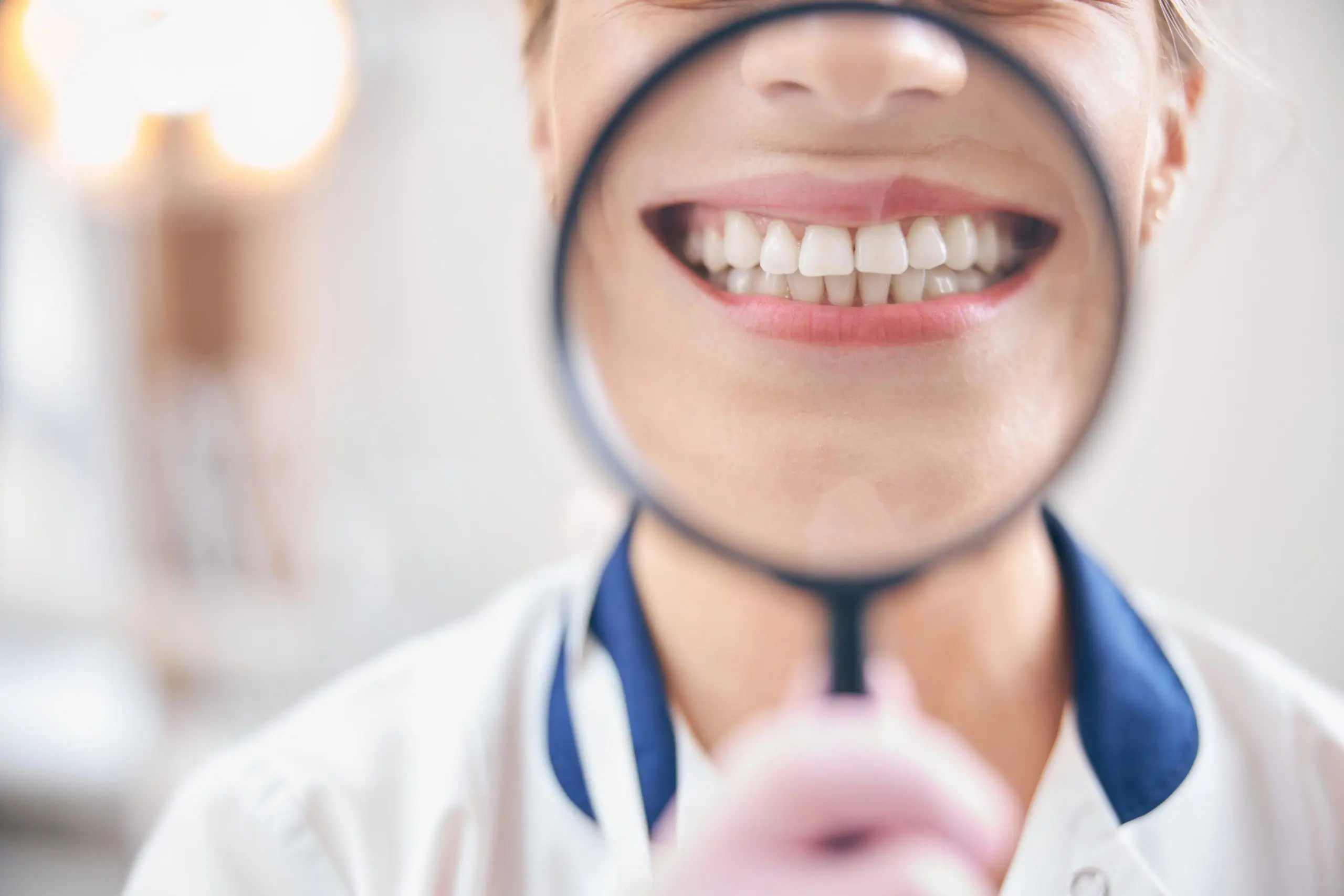 A dental professional looks through a magnifying glass at a smiling person's teeth