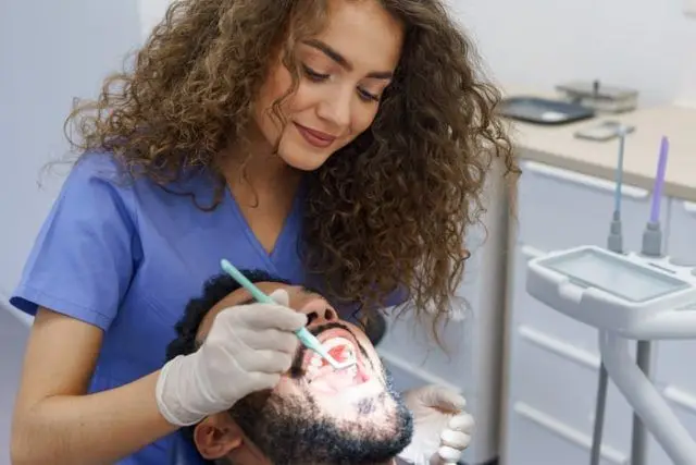 A dentist in purple scrubs examines a smiling patient's teeth with a dental instrument