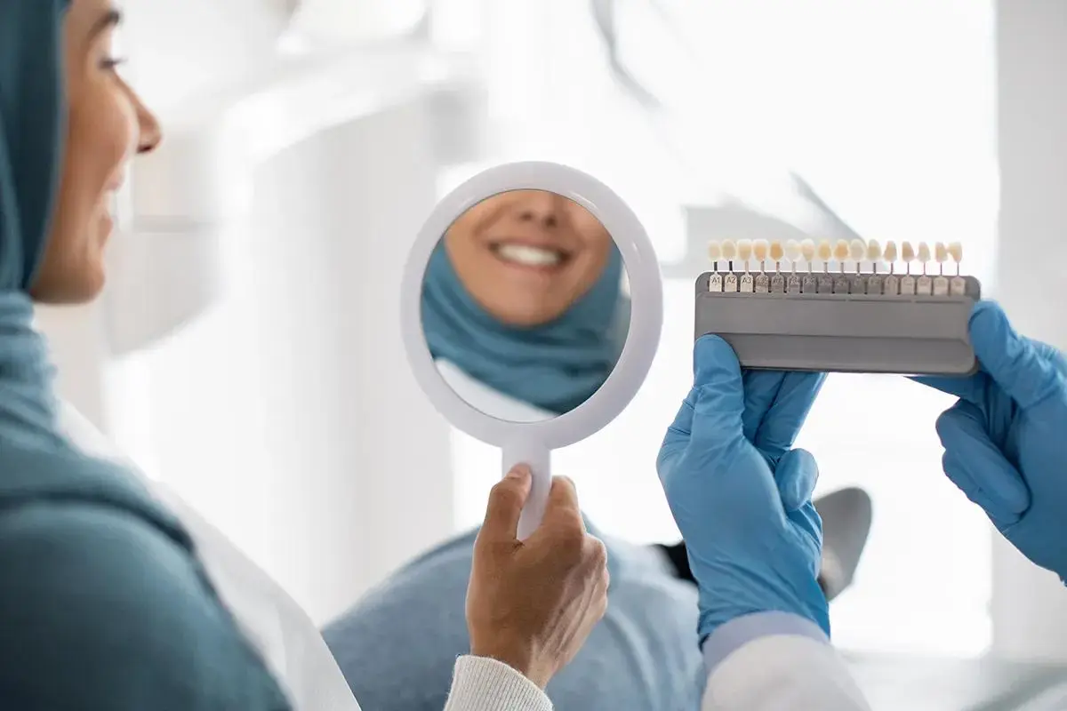 A dental patient smiling in a mirror while a dental professional holds a shade guide to match tooth color.