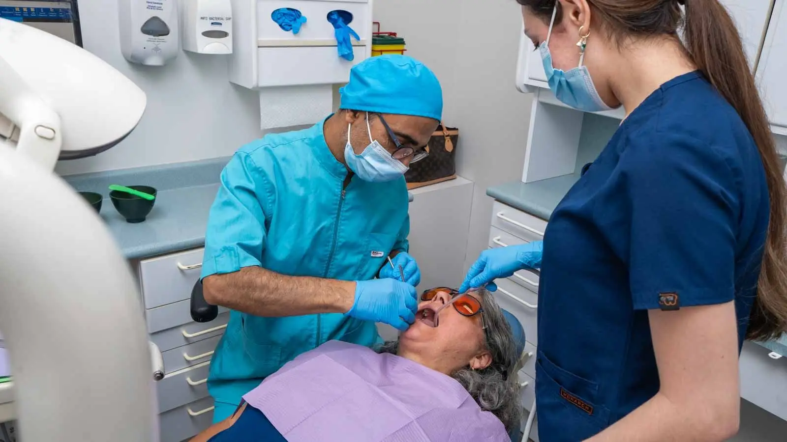 A dentist in blue scrubs treats a reclining patient wearing protective glasses while a dental assistant helps