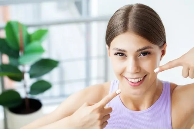 A young woman with straight brown hair and a bright smile is pointing at her teeth. She is wearing a light purple tank top and appears to be indoors, with a potted plant in the blurred background. Her expression is cheerful and she is showcasing her white teeth, possibly indicating satisfaction with dental care or teeth whitening.